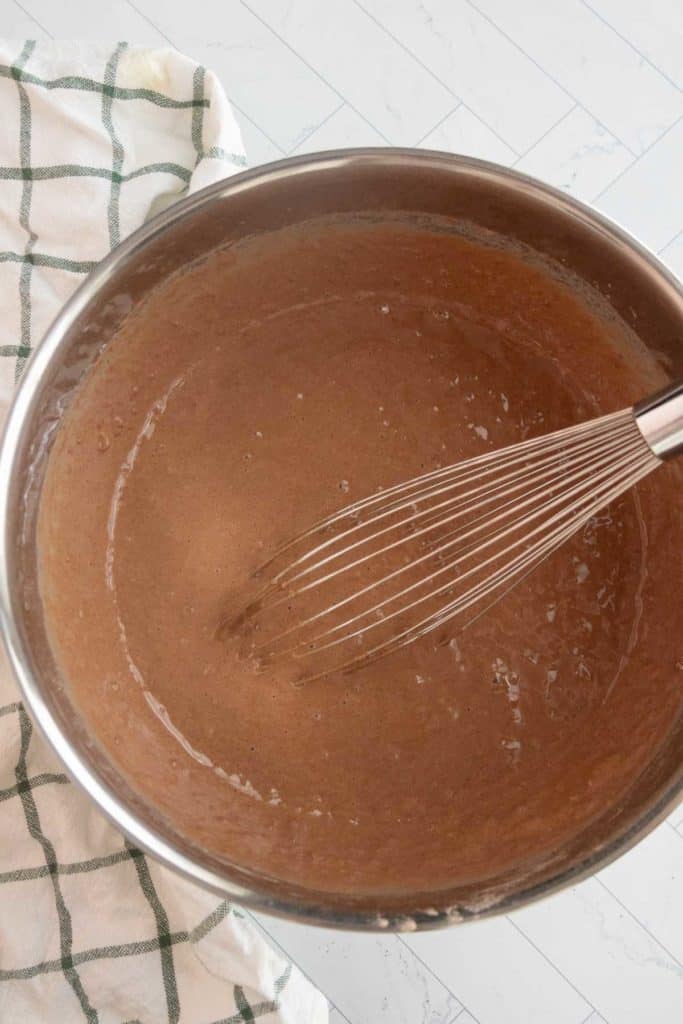 A mixing bowl filled with brown batter and a whisk, placed on a white surface with a green and white checkered cloth nearby.