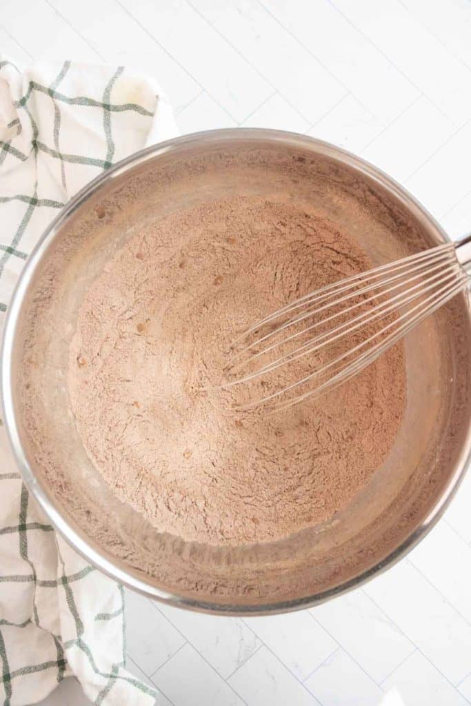 Mixing bowl with whisk and dry brown powder mixture on a light surface, next to a checkered cloth.