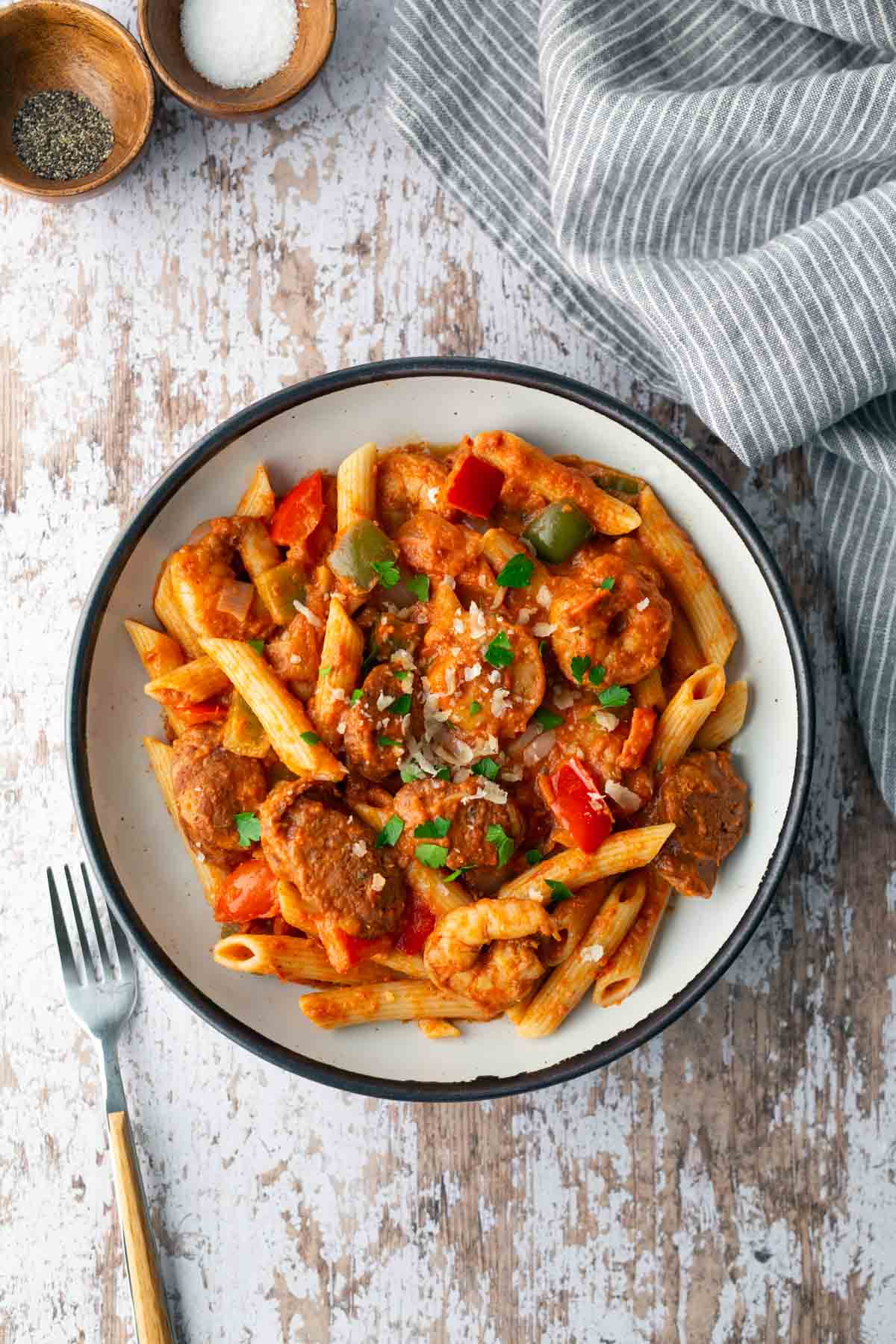 A bowl of penne pasta with meatballs and bell peppers in tomato sauce, topped with parsley and cheese, on a rustic table. A fork, napkin, and small bowls with salt and pepper are nearby.