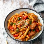 A plate of penne pasta with sausage, bell peppers, and tomato sauce, garnished with parsley, sits on a wooden table next to a fork and striped cloth.