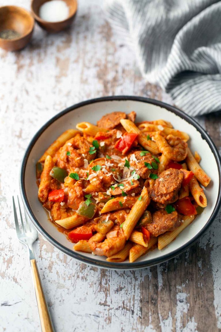 A plate of penne pasta with sausage, bell peppers, and tomato sauce, garnished with parsley, sits on a wooden table next to a fork and striped cloth.