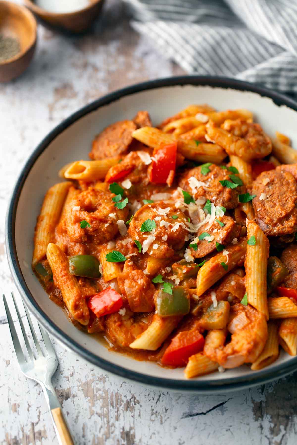 A bowl of penne pasta with chunky tomato sauce, bell peppers, sausage slices, and topped with grated cheese, garnished with parsley. A fork rests beside the bowl on a rustic surface.