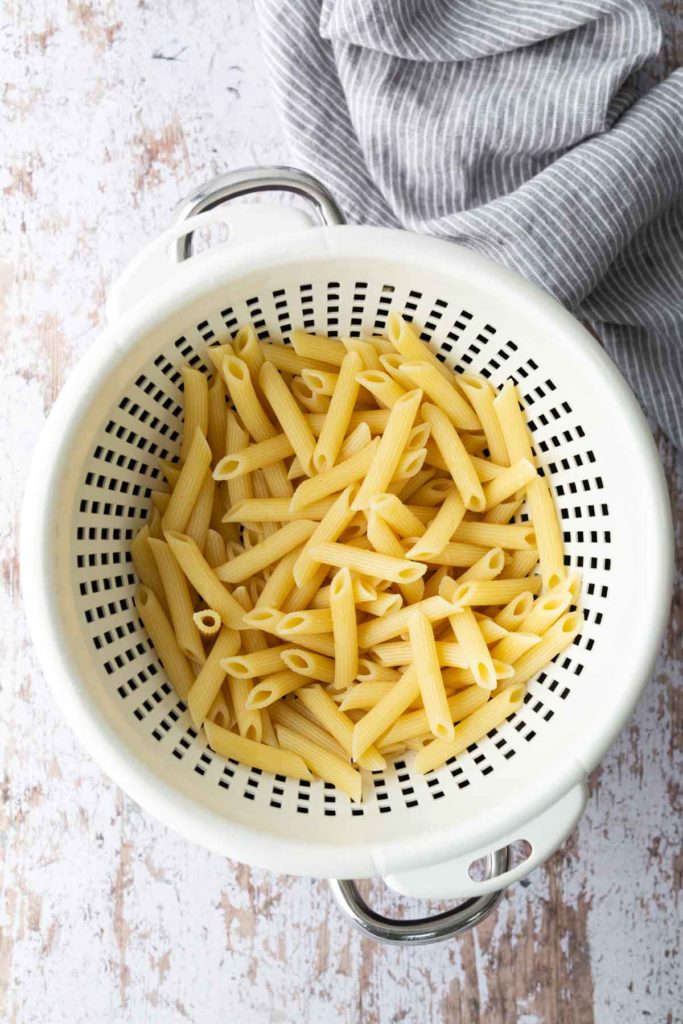 Cooked penne pasta in a white colander on a rustic wooden surface, with a striped cloth nearby.