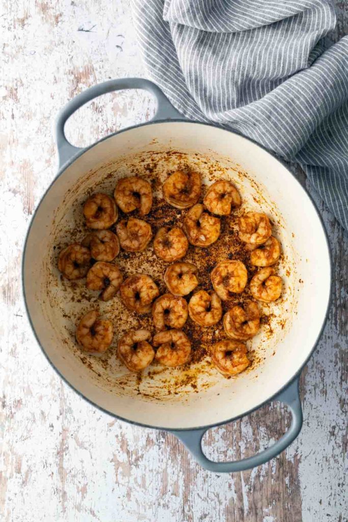 Shrimp seasoned with spices cooking in a large, round, gray pot on a rustic, light-colored surface. A gray-striped cloth is nearby.