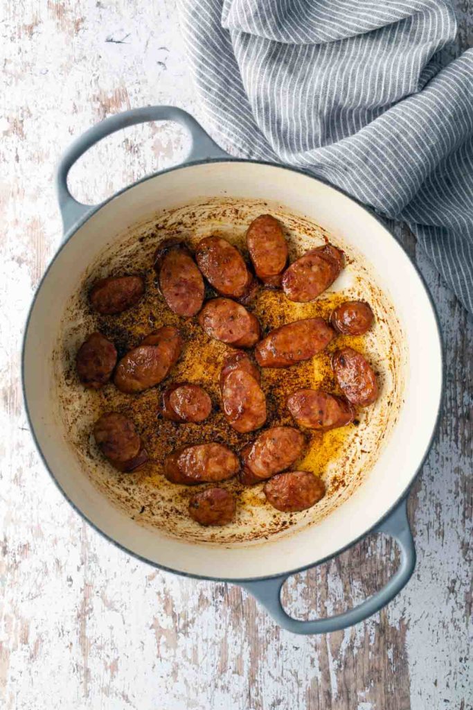 Sliced and cooked sausages in a large pot, with oil and spices, on a textured surface next to a striped cloth.