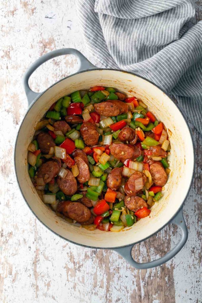 A pot filled with cooked sausages, diced bell peppers, and onions. The pot is on a textured white surface with a striped cloth nearby.