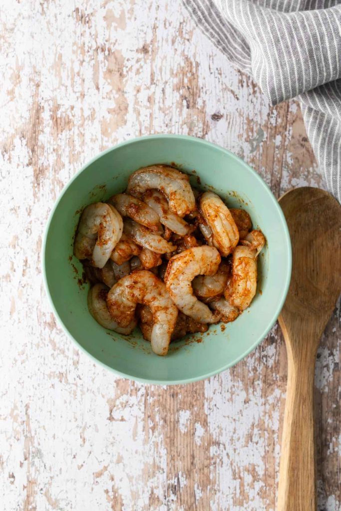 Bowl of seasoned raw shrimp on a worn wooden surface with a wooden spoon beside it.