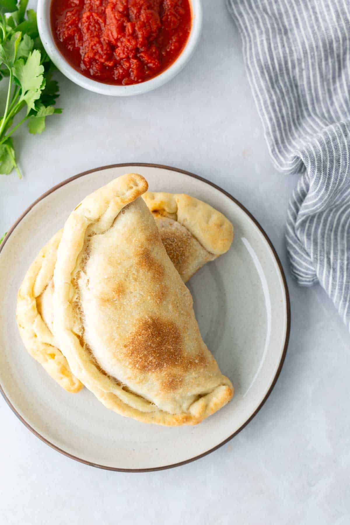 Two baked calzones on a plate with a side of red sauce, parsley leaves, and a striped cloth nearby.