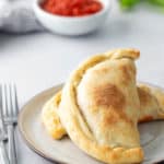 A plate with two baked calzones on it. A bowl of red sauce, a striped cloth, and parsley are blurred in the background.