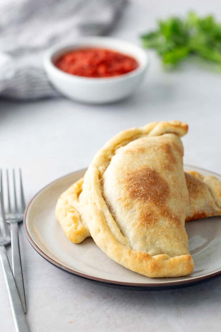 A plate with two baked calzones on it. A bowl of red sauce, a striped cloth, and parsley are blurred in the background.