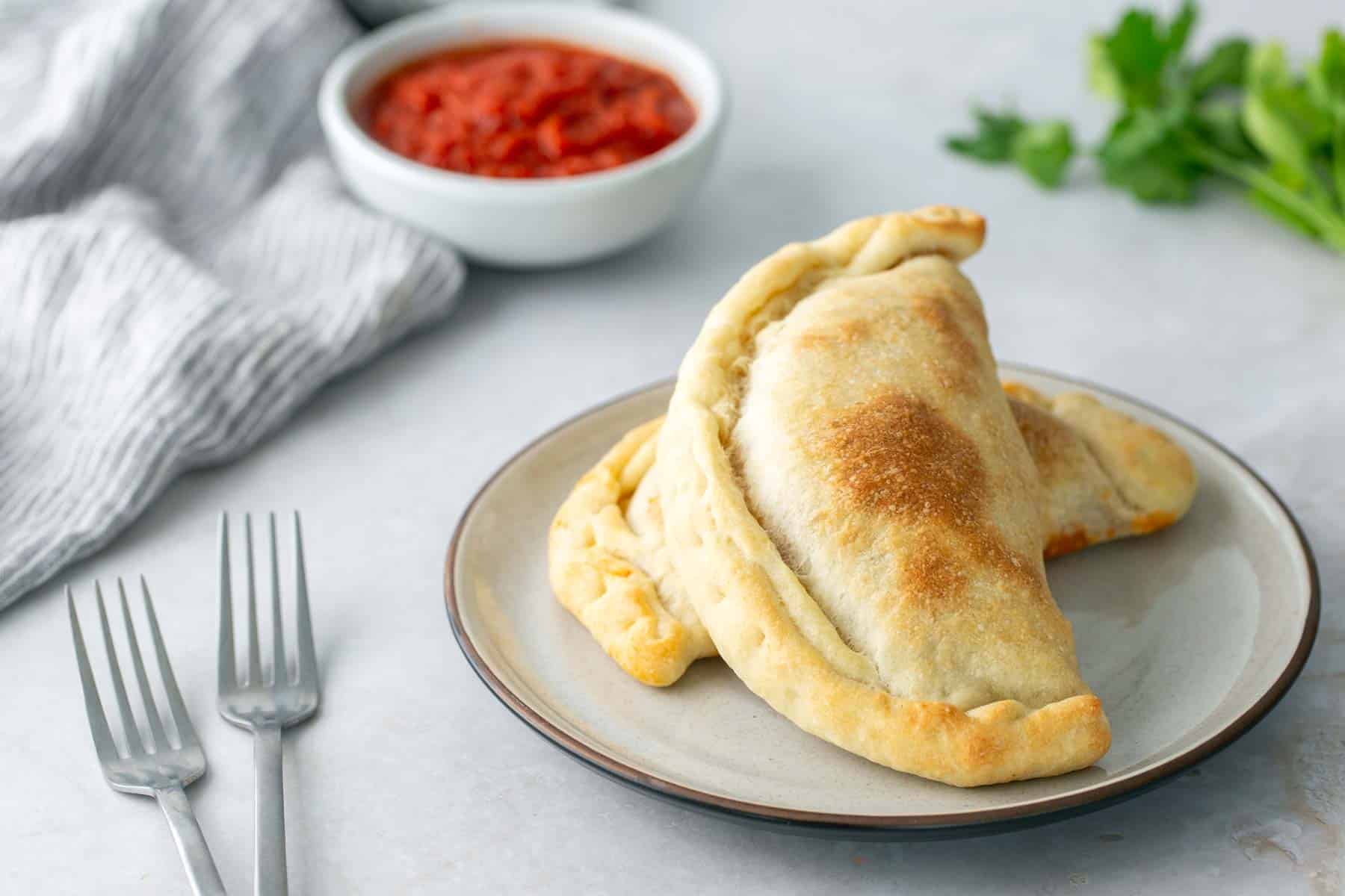 Two baked calzones on a plate beside marinara sauce, with two forks and a striped cloth nearby.