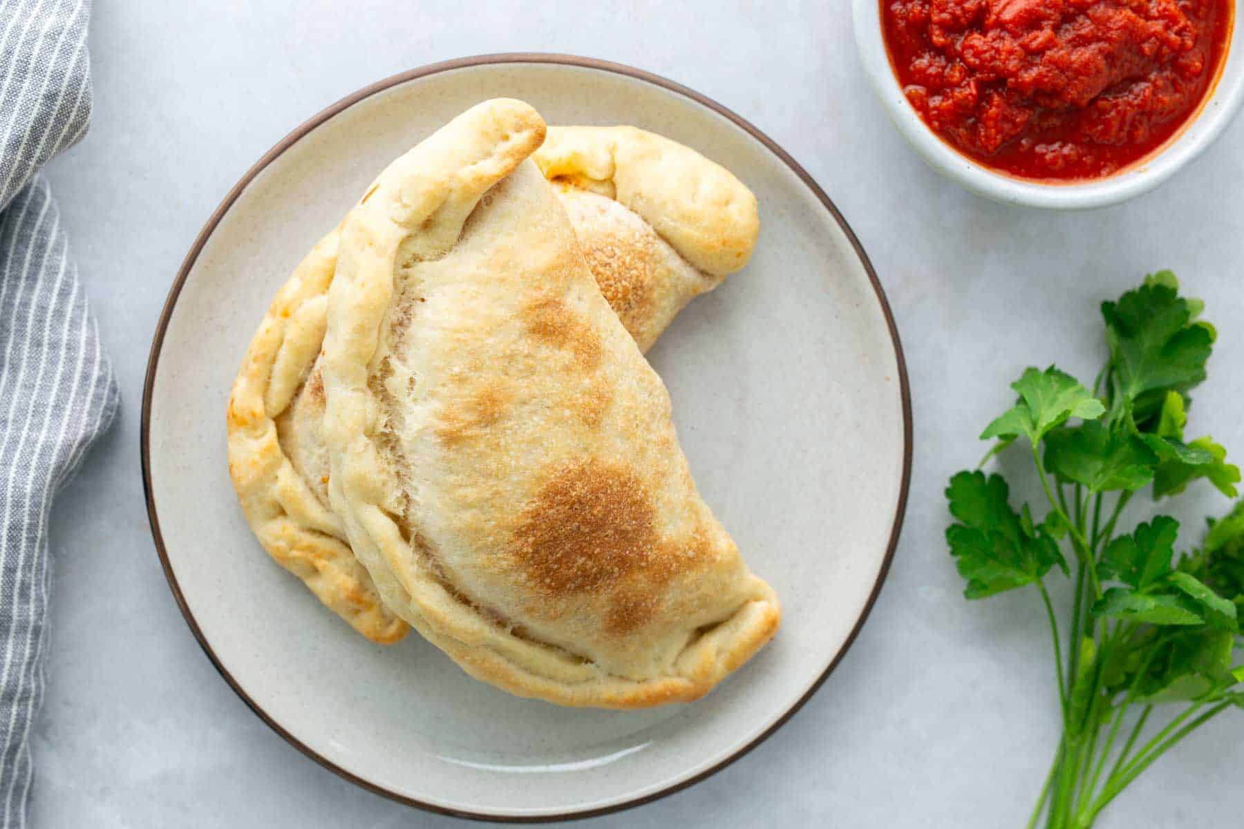 Two baked calzones on a plate with a small bowl of red sauce and fresh parsley beside them.