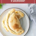 Two pepperoni calzones on a plate, garnished with parsley, with a side of red sauce and a striped cloth nearby.