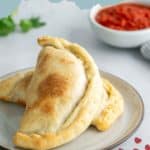 A plate with a baked pepperoni calzone. In the background, there's a bowl of tomato sauce and some fresh herbs.