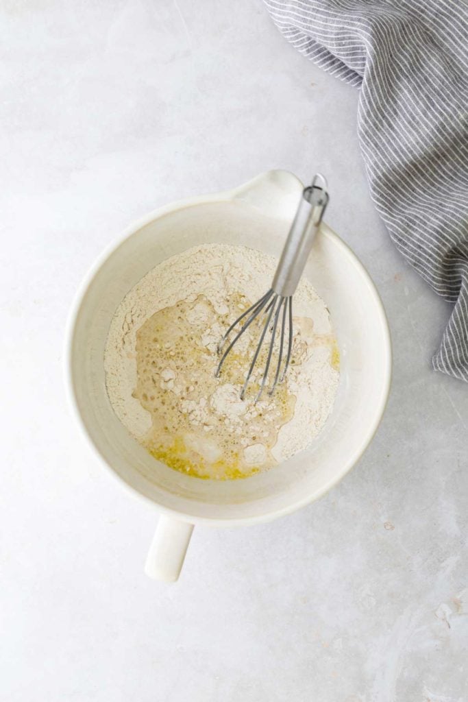 A ceramic bowl with flour, wet ingredients, and a whisk on a light surface. A striped cloth is partially visible on the side.