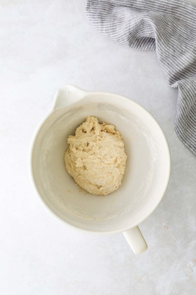 A dough ball in a white mixing bowl on a light surface with a gray striped cloth nearby.