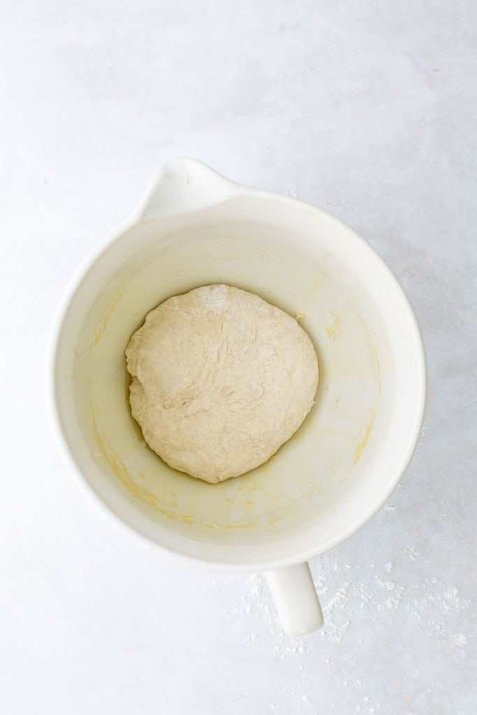 A ball of dough in a white mixing bowl on a light surface.