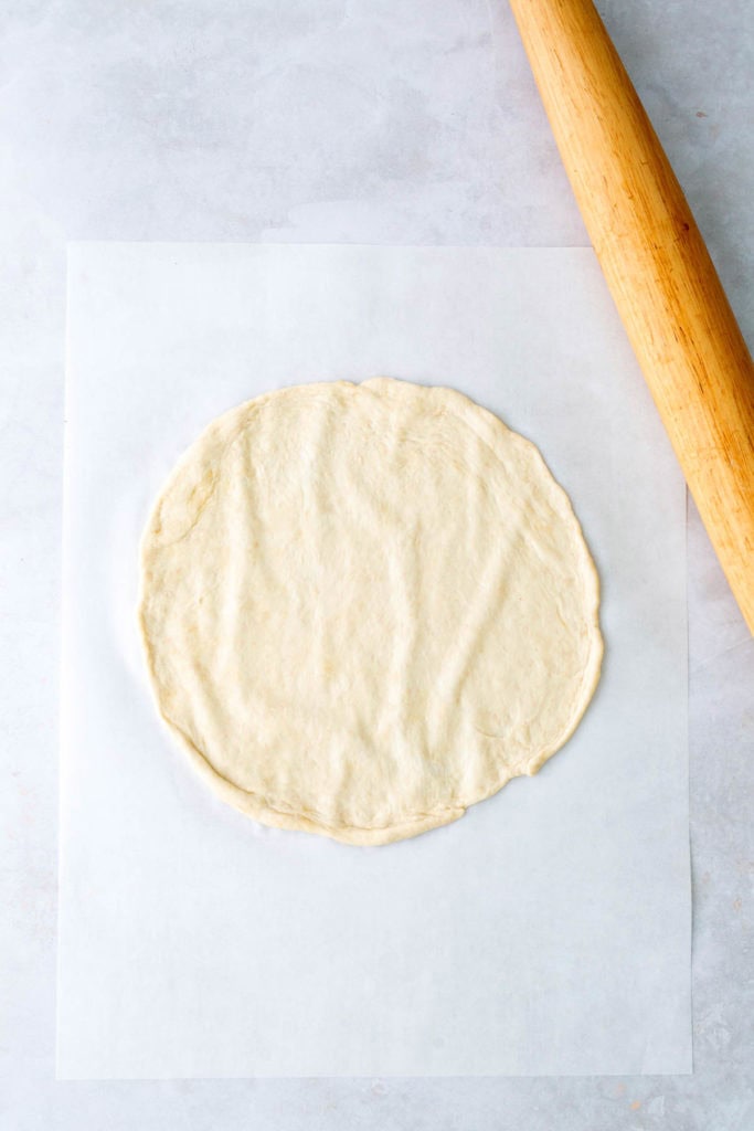 Round pizza dough on parchment paper next to a wooden rolling pin on a light surface.