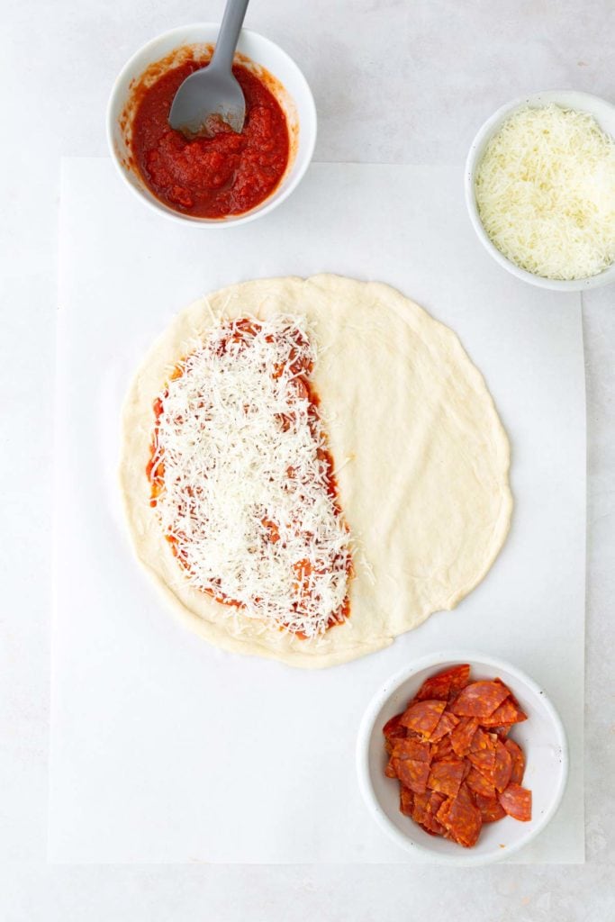 Pizza dough half-covered with tomato sauce and shredded cheese, surrounded by bowls of sauce, cheese, and pepperoni.