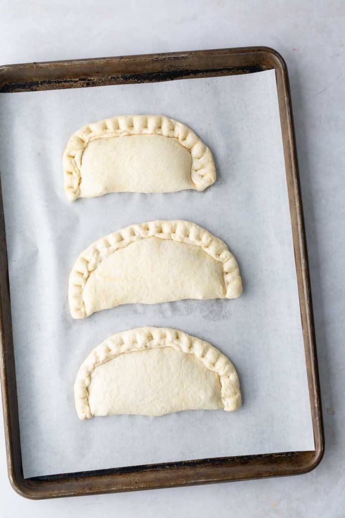 Three uncooked empanadas with crimped edges on a parchment-lined baking sheet.