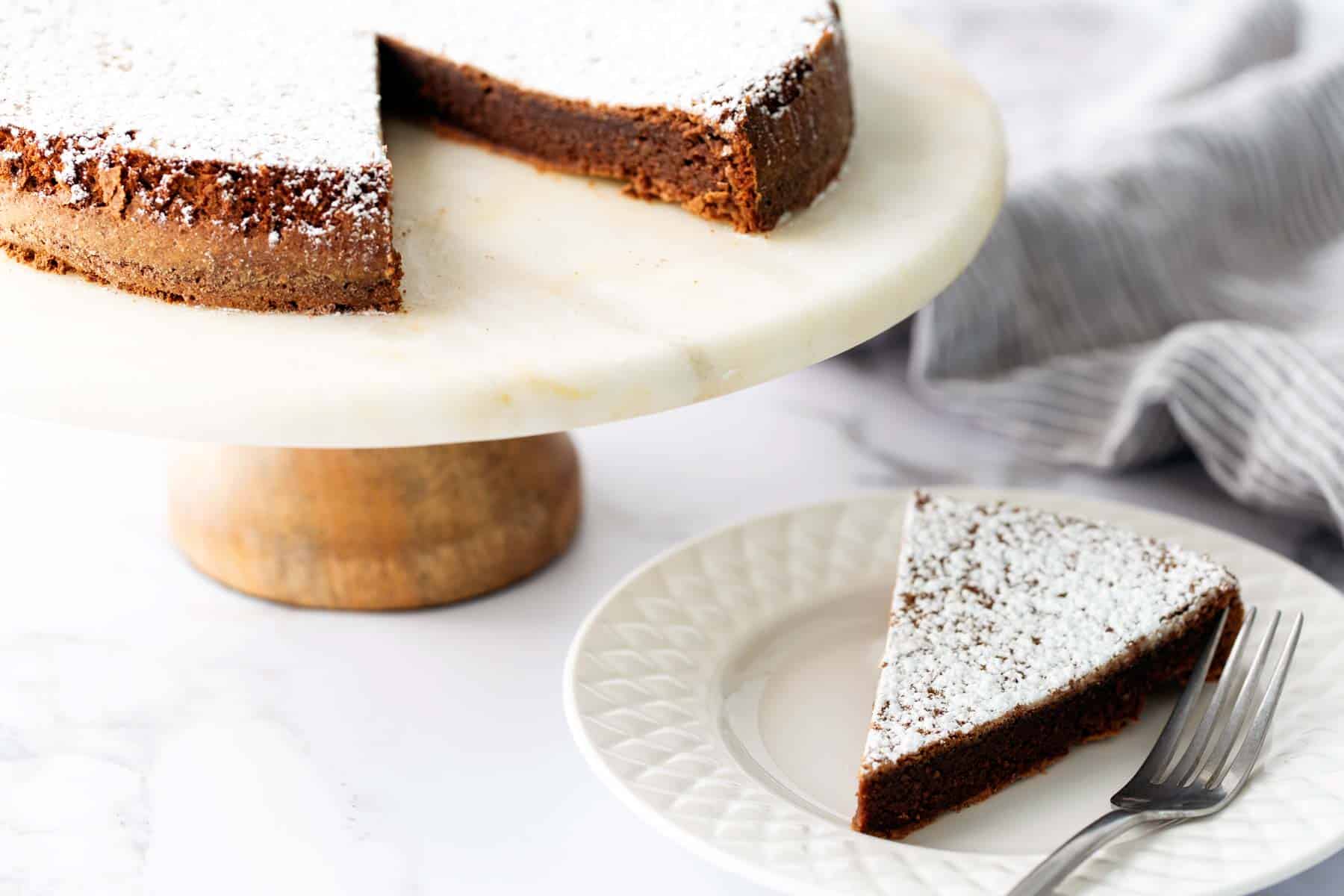 A chocolate cake with powdered sugar on top is on a marble cake stand. A slice is cut and placed on a white plate with a fork beside it.