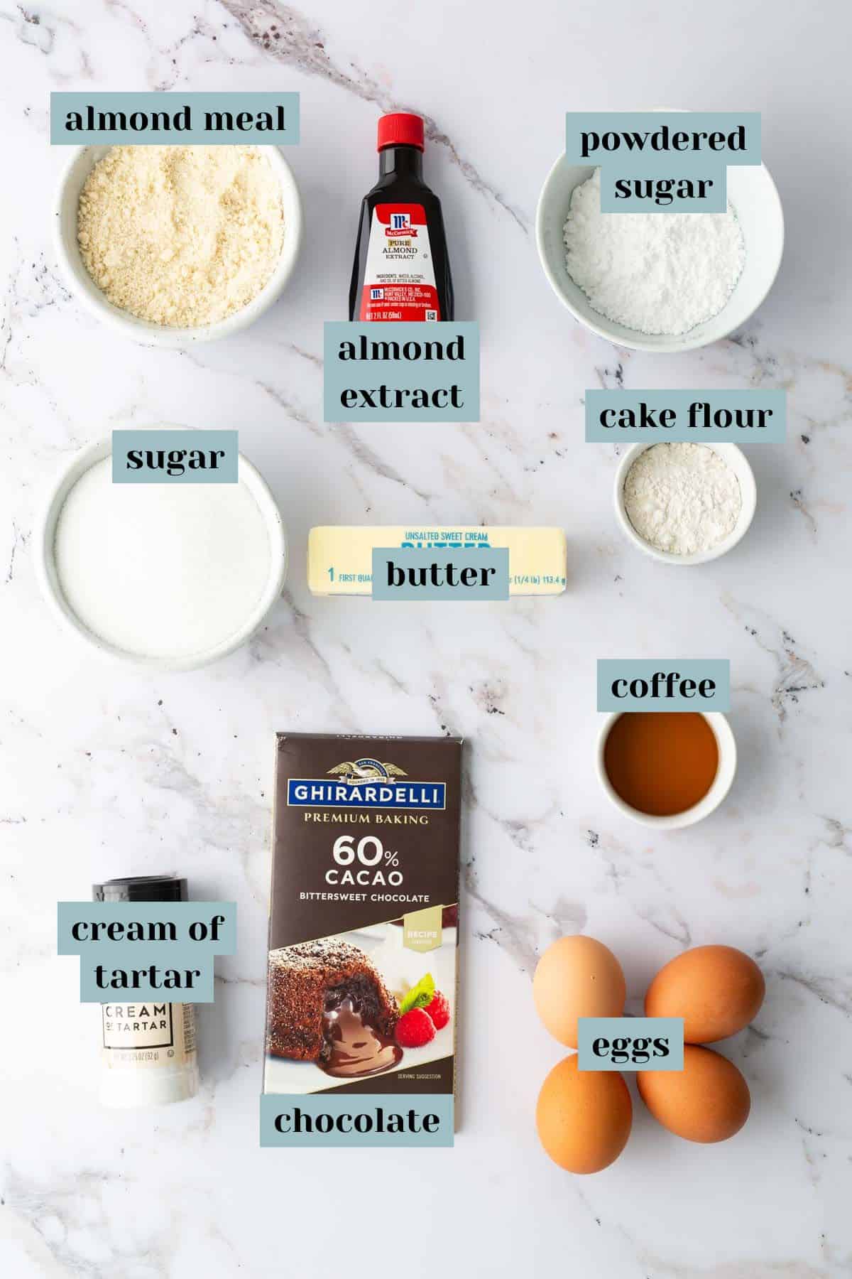 Ingredients arranged on a marble surface: almond meal, powdered sugar, sugar, almond extract, cake flour, butter, coffee, eggs, cream of tartar, and a chocolate bar.