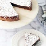 A chocolate cake dusted with powdered sugar sits on a marble stand. One slice is cut and placed on a white plate with a fork on the side.