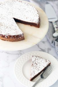 A chocolate cake dusted with powdered sugar sits on a marble stand. One slice is cut and placed on a white plate with a fork on the side.