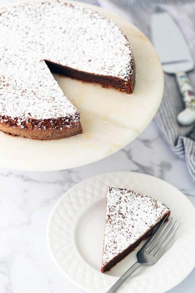 A chocolate cake dusted with powdered sugar sits on a marble stand. One slice is cut and placed on a white plate with a fork on the side.