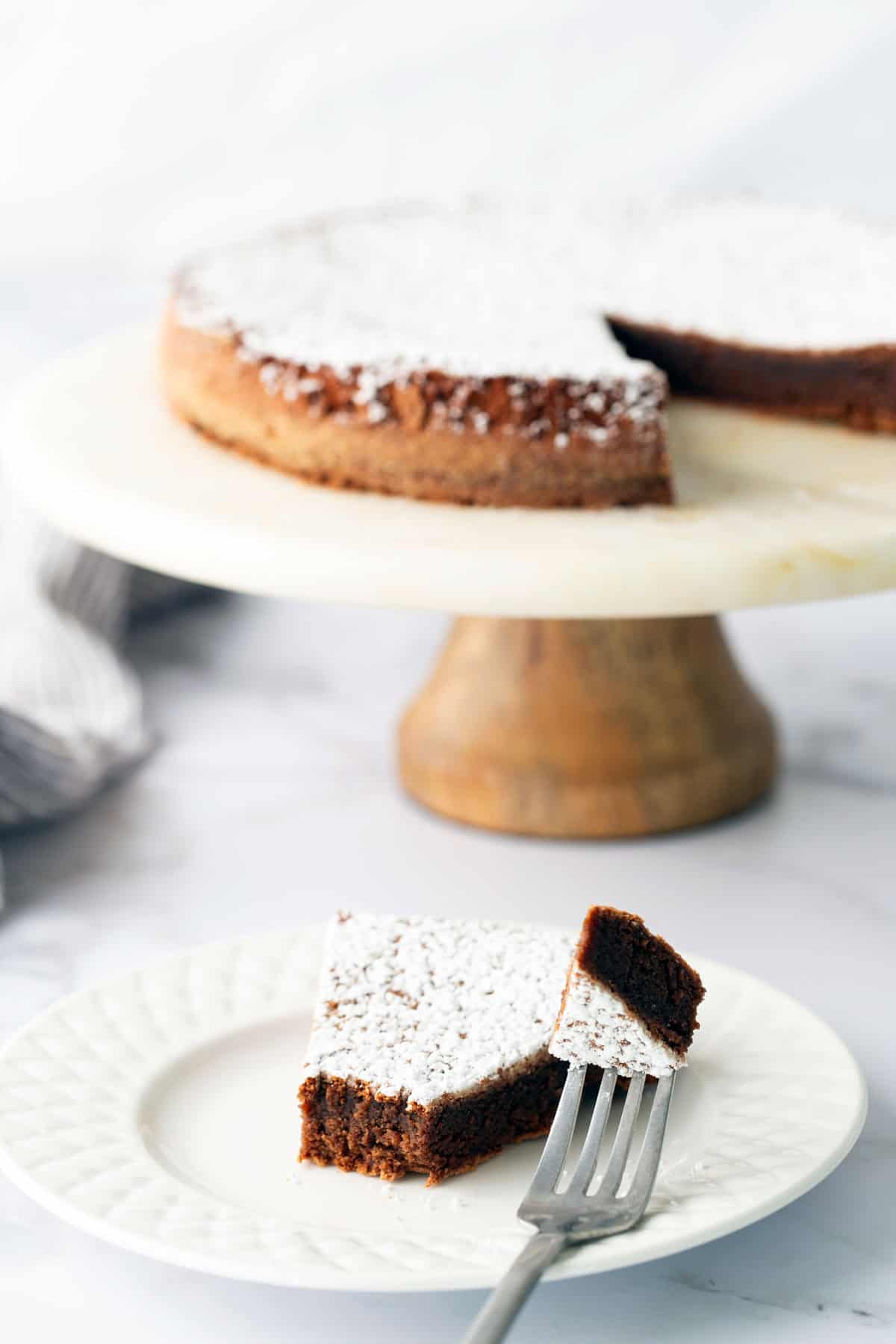 Slice of chocolate cake on a plate with a fork, whole cake in the background on a wooden stand. Both topped with powdered sugar.