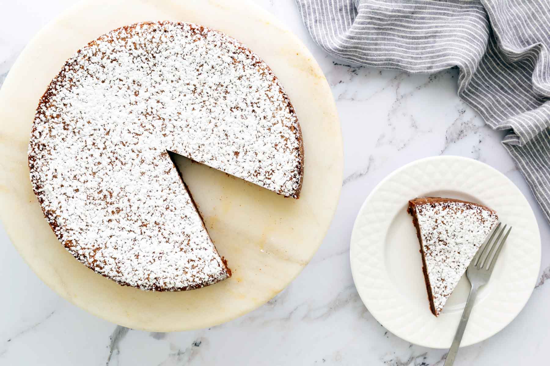 A round cake dusted with powdered sugar, missing one slice. The slice is on a plate with a fork beside it. A striped cloth is nearby on a marble surface.