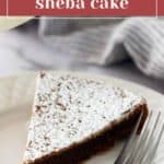 A slice of Queen of Sheba cake is on a white plate, dusted with powdered sugar, with a fork beside it. A striped cloth is in the background.