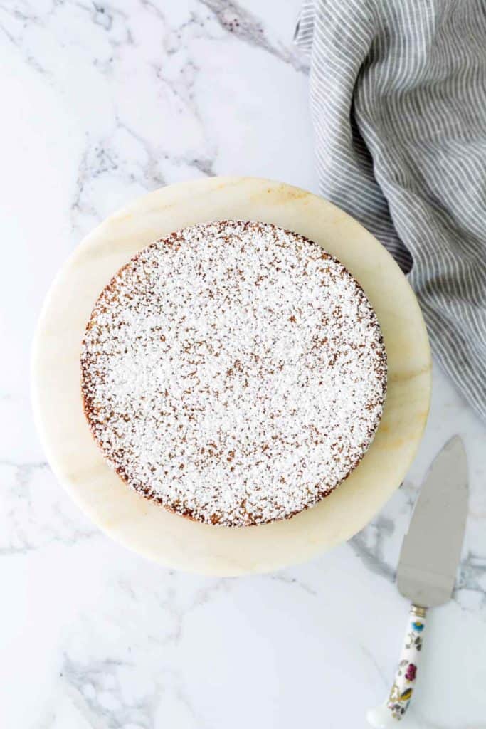 A round cake topped with powdered sugar on a marble surface, next to a serrated knife with a decorative handle.