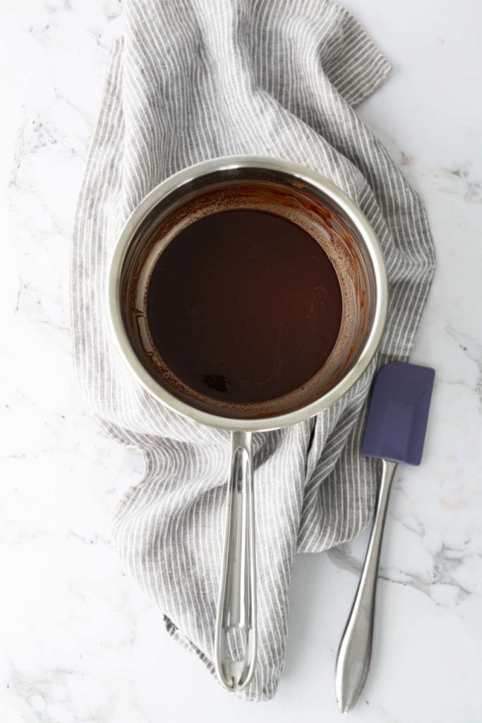 A saucepan filled with melted chocolate sits on a gray striped cloth. A blue spatula lies next to the pan on a marble surface.