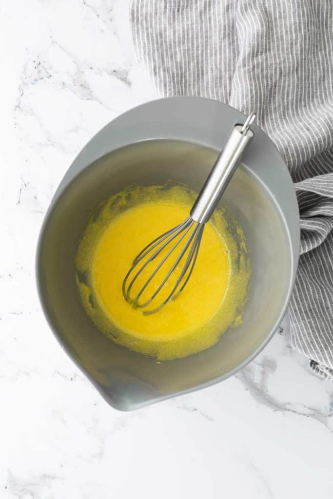 A grey bowl with a wire whisk containing a yellow liquid mixture on a marble surface next to a striped fabric.