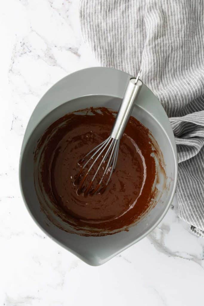 A gray bowl filled with chocolate batter and a whisk on a marble countertop next to a striped cloth.