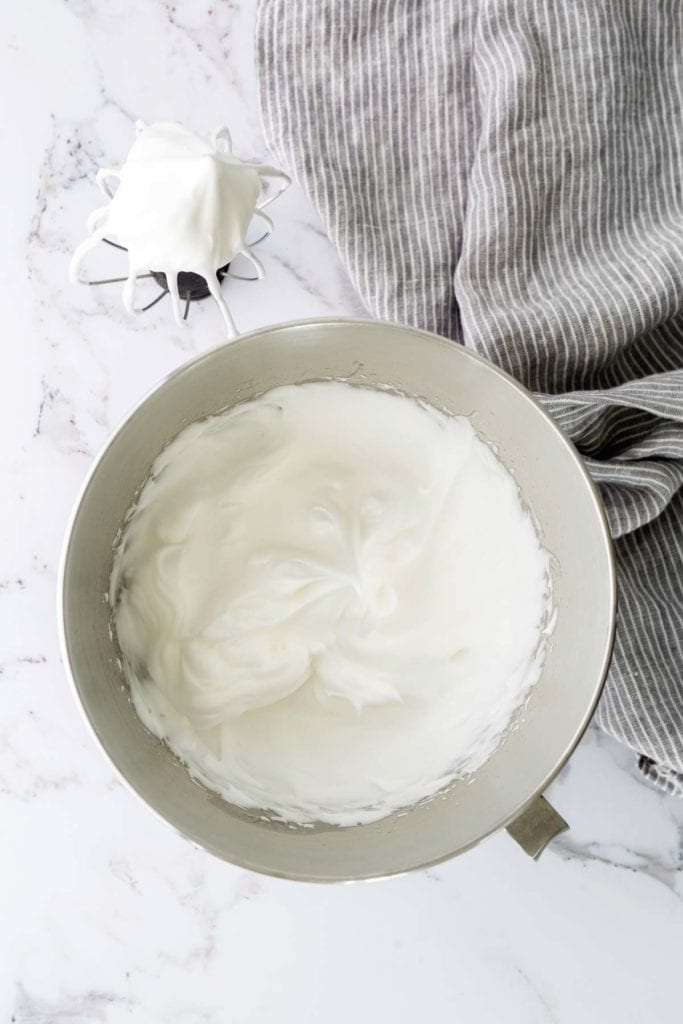 Bowl of whipped cream next to a whisk with cream on it on a marble surface. Striped cloth to the right.