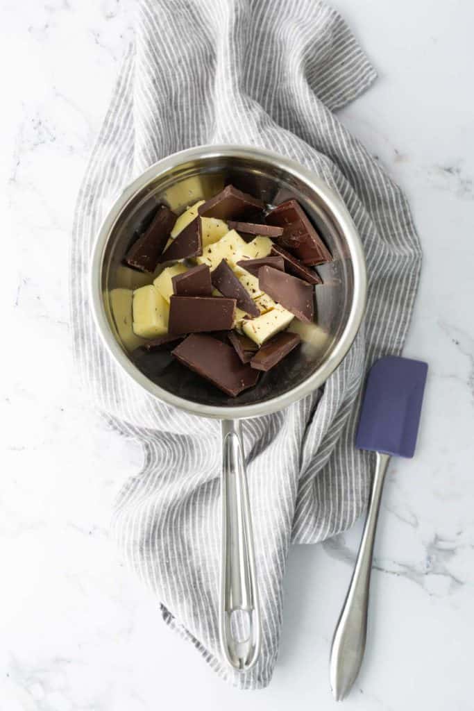 A saucepan filled with dark and white chocolate pieces, placed on a gray striped cloth with a blue spatula beside it.