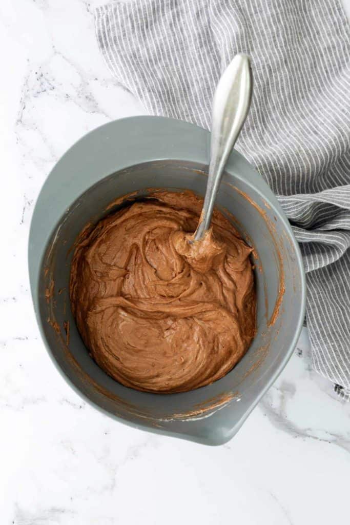 A gray bowl with chocolate batter and a spoon on a marble surface, next to a striped cloth.