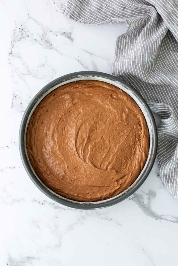 Round pan filled with smooth chocolate batter on a marble surface. A gray striped cloth is beside the pan.