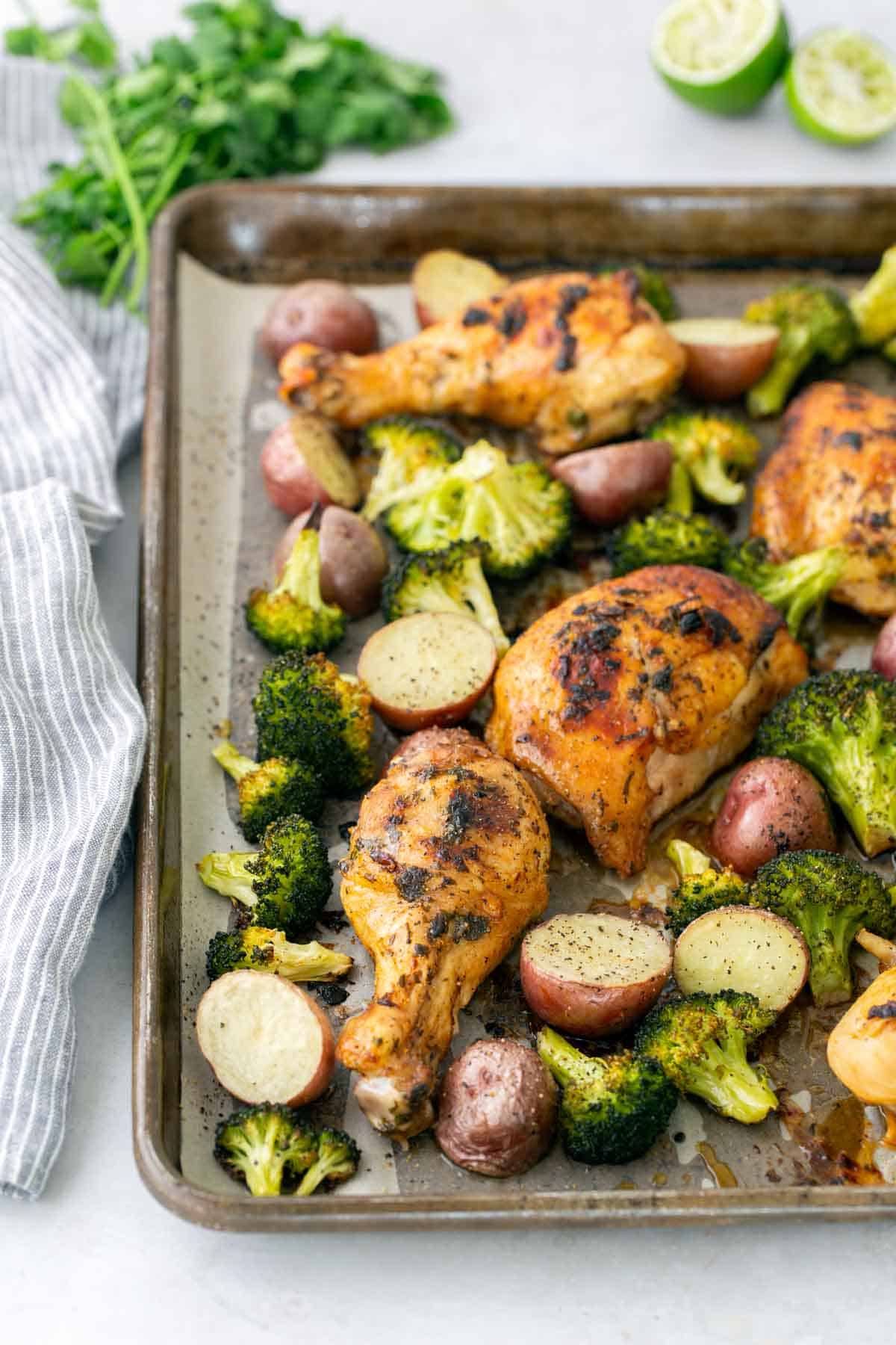 Sheet pan with roasted chicken drumsticks, broccoli, and red potatoes. Striped napkin and lime wedges nearby.