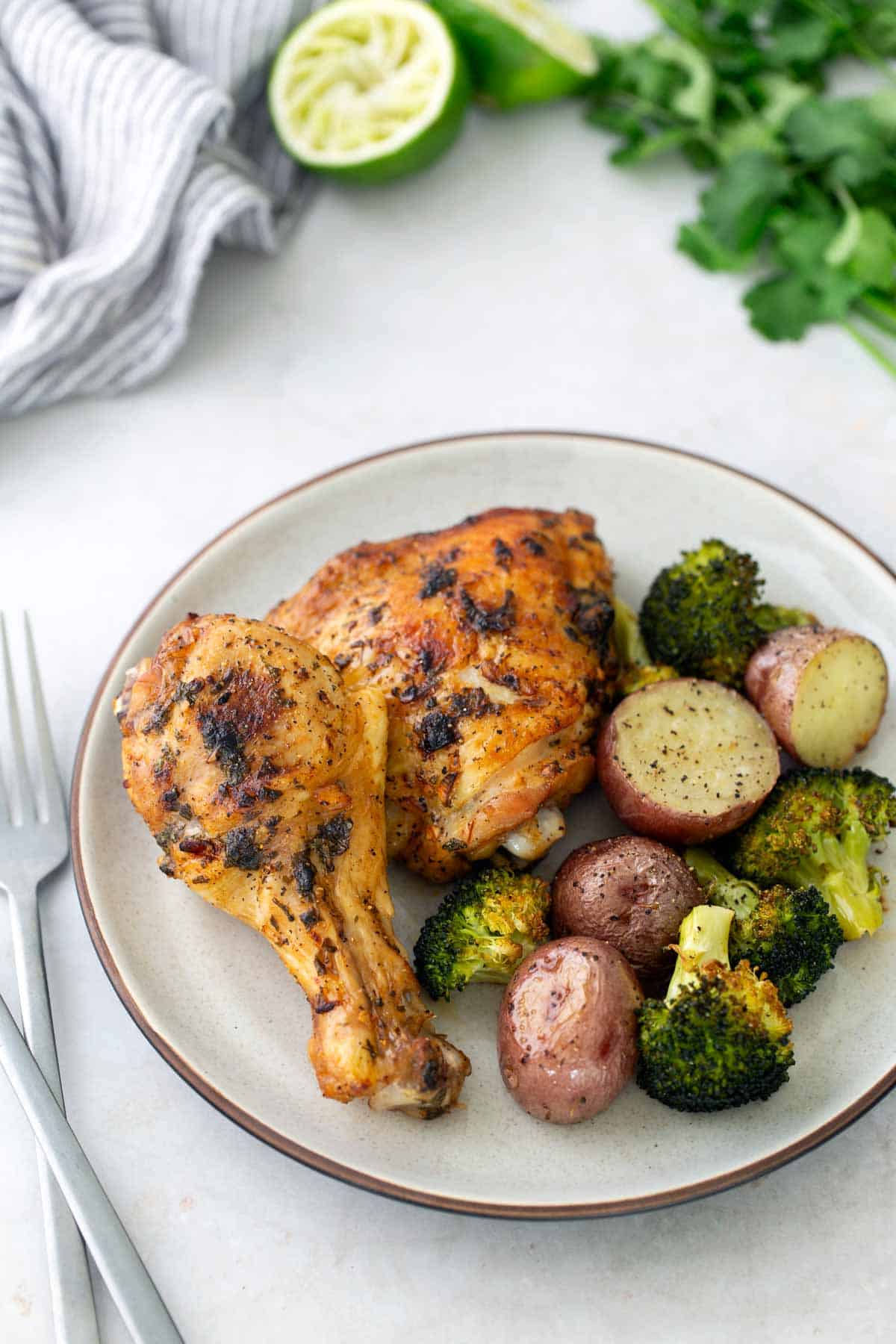 Plate with roasted chicken drumstick and thigh, seasoned potatoes, and broccoli. Lime half and napkin in the background.