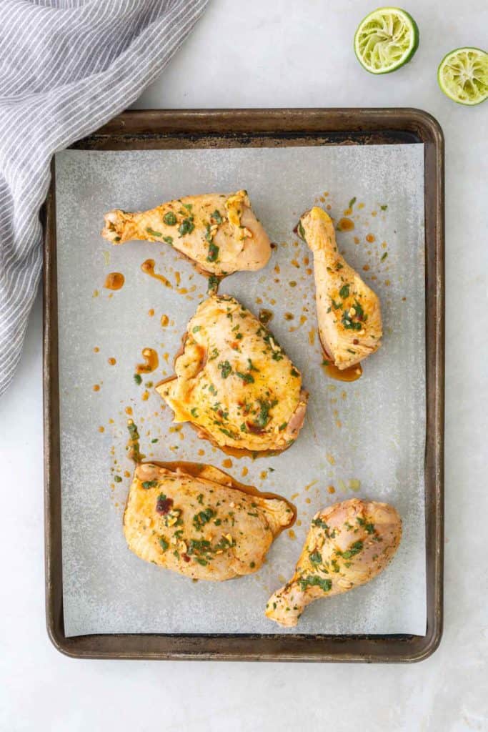 Baked seasoned chicken pieces on a parchment-lined tray, garnished with herbs. Two squeezed lime halves are visible in the top corner. A striped cloth is on the side.