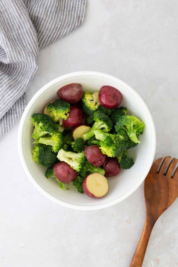 A bowl with broccoli florets and red potatoes on a light surface. A wooden utensil and a striped cloth are nearby.