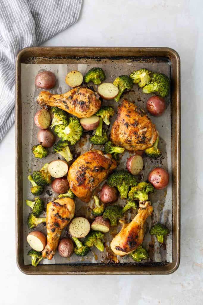 Baked chicken drumsticks and thighs with broccoli and halved red potatoes on a sheet pan.