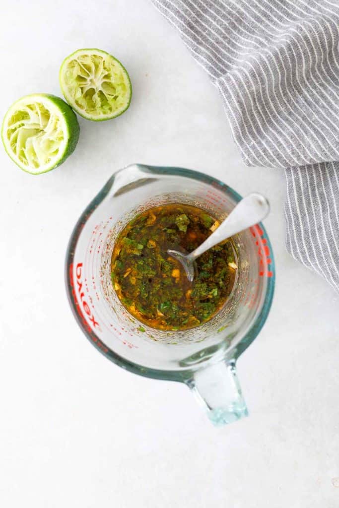 A measuring cup with mixed marinade, a spoon inside, next to squeezed lime halves and a gray striped cloth on a white surface.