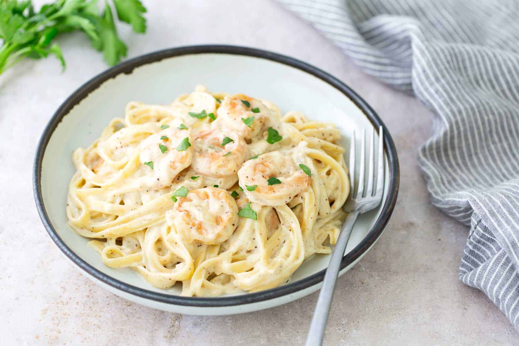 A bowl of fettuccine pasta with creamy sauce and shrimp, garnished with parsley. A fork rests on the bowl's rim. A gray-striped cloth napkin is beside it.