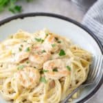 A plate of creamy fettuccine pasta topped with shrimp and garnished with parsley. A fork rests on the side of the plate. A bunch of parsley and a striped cloth are in the background.