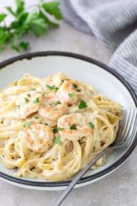 A plate of creamy fettuccine pasta topped with shrimp and garnished with parsley. A fork rests on the side of the plate. A bunch of parsley and a striped cloth are in the background.