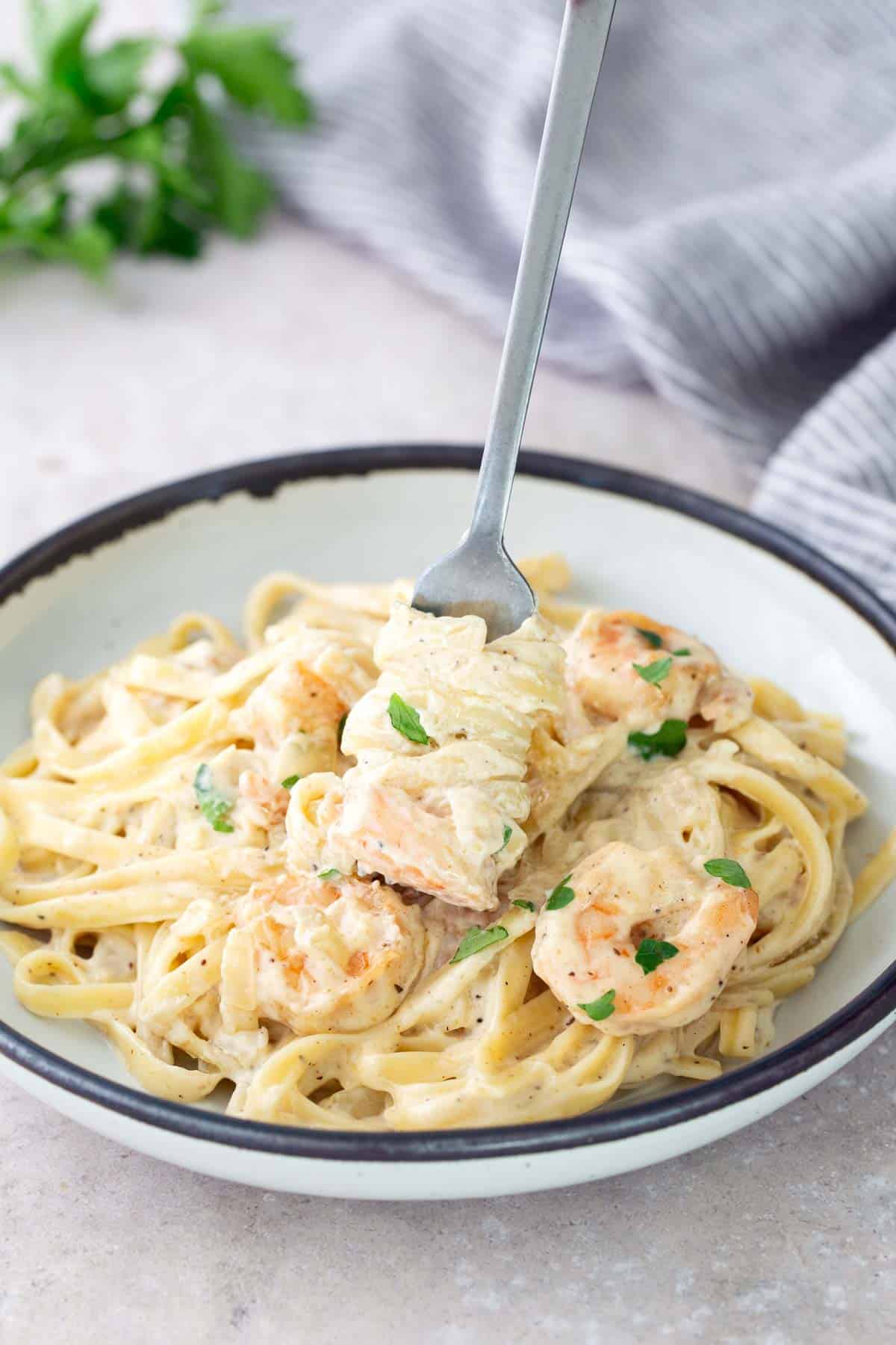 A bowl of fettuccine pasta with creamy sauce, topped with shrimp and garnished with parsley. A fork is twirling pasta above the bowl.